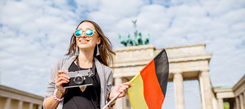 Uśmiechnięta dziewczyna w okularach przeciwsłonecznych trzyma w rękach flagę Niemiec i aparat, w tle Brama Brandenburska 