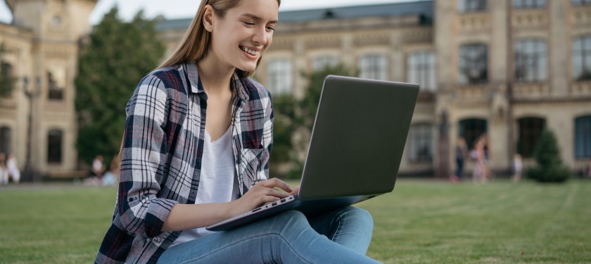 Młoda dziewczyna siedzi na trawniku przed uczelnią, trzyma na kolanach laptop i uśmiecha się do siebie