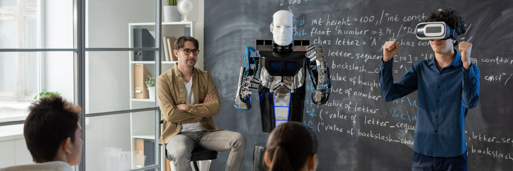 Chłopak w okularach VR i robot stoją przed kredową tablicą. Obok siedzi nauczyciel i przygląda się