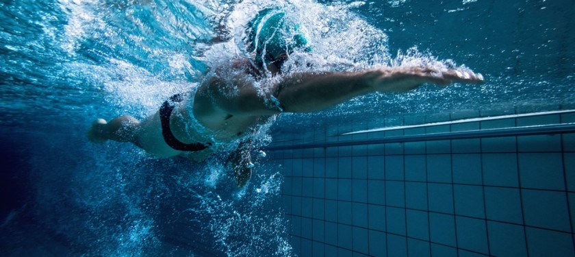 Sportowiec dynamicznie płynący pod wodą w basenie, woda jest wzburzona, całość w niebiesko-białej kolorystyce
