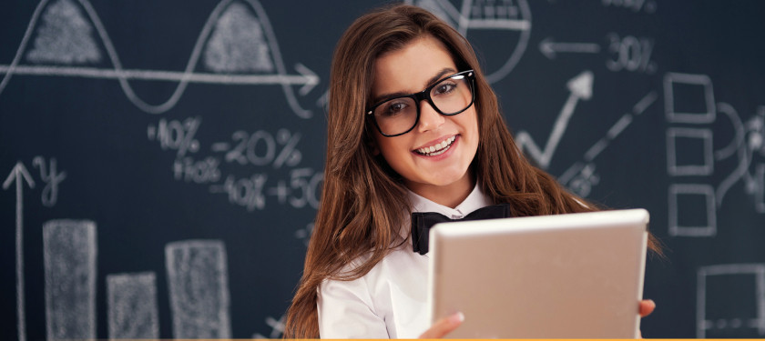 Dziewczyna w ciemnych włosach i okularach stoi przed tablicą z matematycznymi wzorami, a w ręku trzyma teczkę