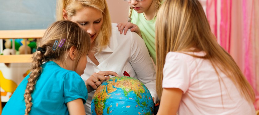 Dzieci w wieku wczesnoszkolnym wraz z nauczycielką siedzą wokół globusu, pani nauczycielka pokazuje palcem na globus